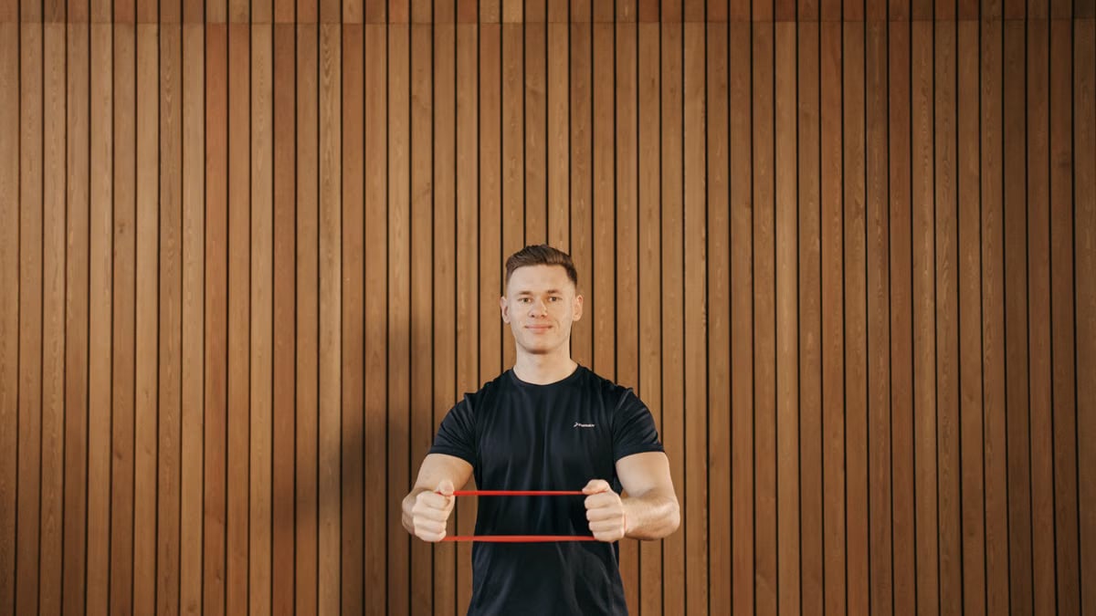 Man in a black shirt using a resistance band for exercise against a wooden wall indoors.