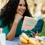 Smiling woman reading a book at a picnic in a sunny park setting.